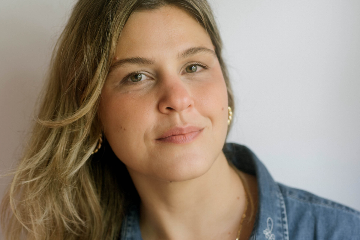 Mulher jovem de cabelos loiros e ondulados, com olhos verdes e sorriso discreto, usando brincos dourados e camisa jeans, em fundo branco