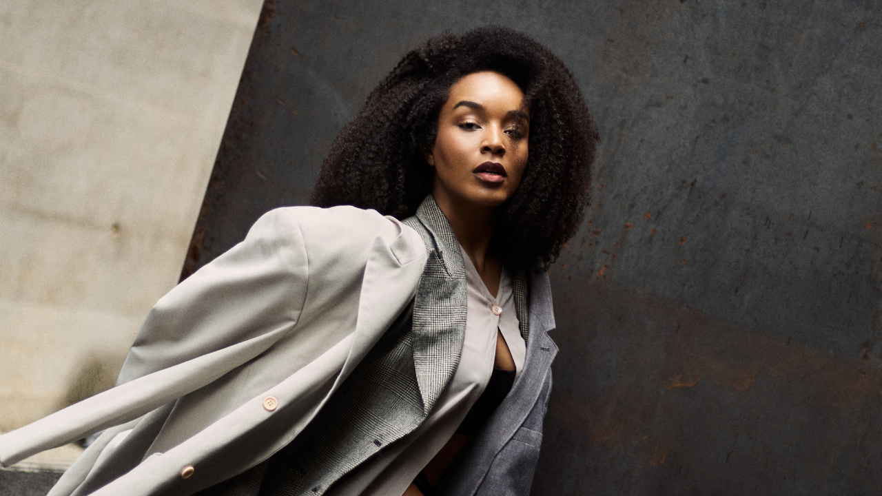 Mulher negra com cabelo crespo volumoso, vestindo blazer cinza claro sobre blazer xadrez e camisa aberta, olhando diretamente para a câmera com expressão séria, em fundo escuro e texturizado
