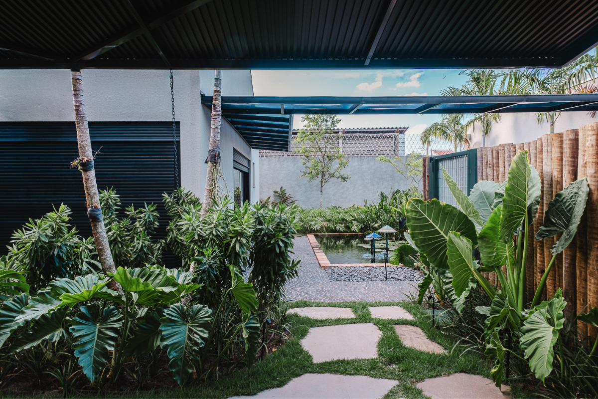 Um jardim exuberante com um caminho de pedras irregulares sobre a grama, ladeado por folhagens verdes. Ao fundo, um lago ornamental com pedras escuras e uma fonte, cercado por plantas. À direita, um muro de madeira vertical e, à esquerda, a parede de uma casa com persianas escuras. O céu azul com nuvens brancas é visível acima de um telhado metálico