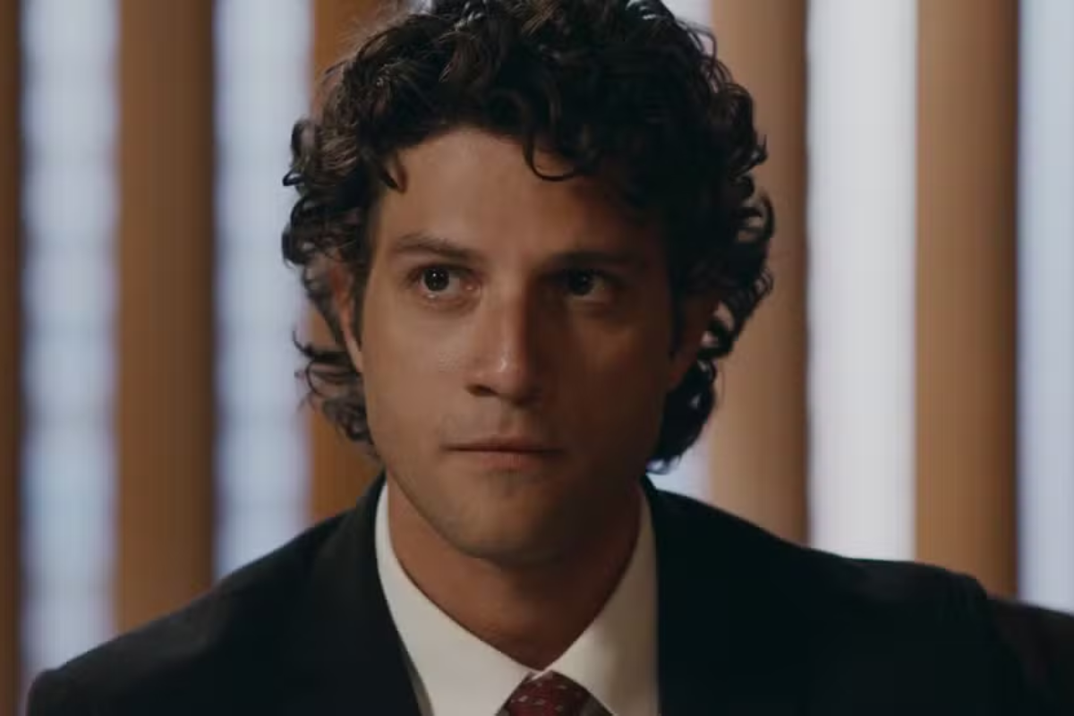 Homem jovem de pele clara, cabelo castanho escuro encaracolado, vestindo terno preto, camisa branca e gravata bordô, olhando para a direita com expressão séria