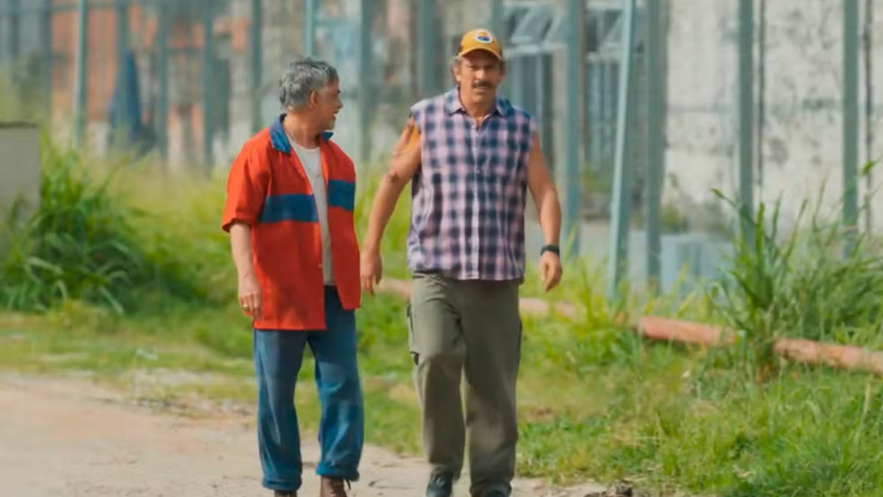 Dois homens caminham em uma estrada de terra. O da esquerda, com cabelo grisalho, usa camisa vermelha com listras azuis e calça jeans. O da direita, de bigode e boné amarelo, veste camisa xadrez sem mangas e calça cargo verde. Ambos parecem conversar em um ambiente externo com vegetação e estruturas ao fundo