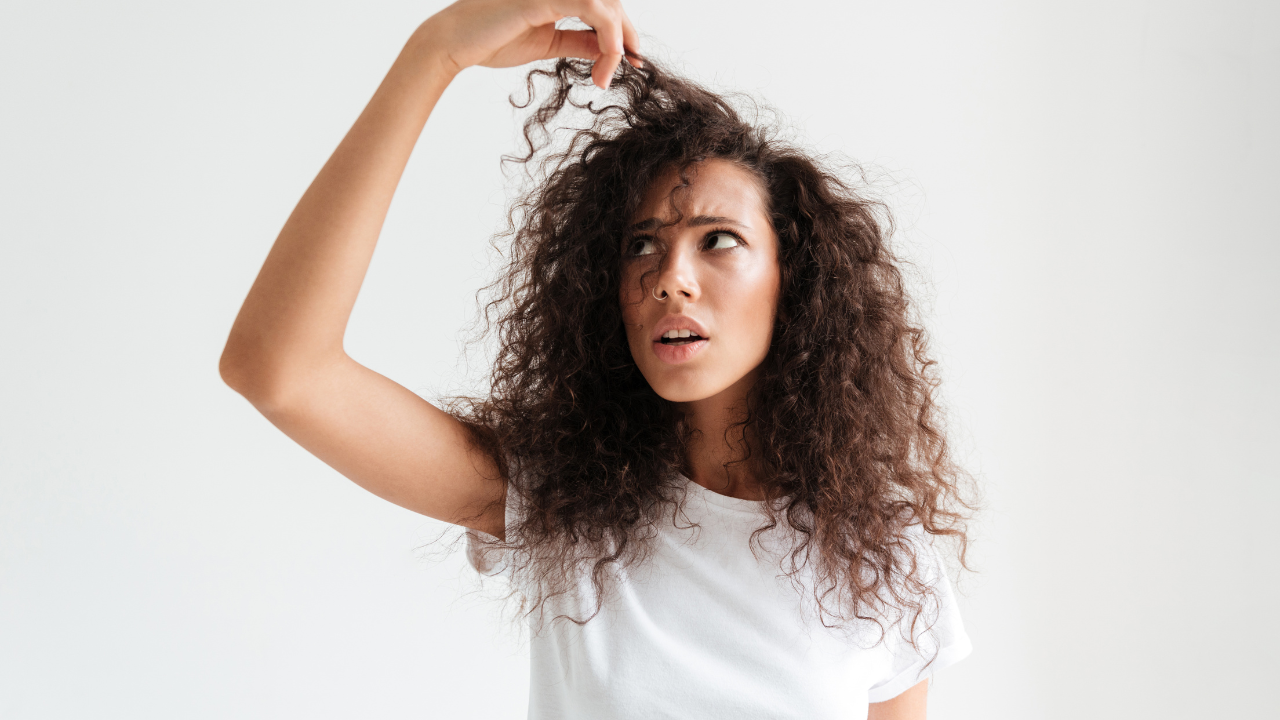 Mulher jovem de pele morena e cabelos cacheados escuros, vestindo camiseta branca, com expressão de surpresa e preocupação, olhando para cima enquanto segura uma mecha de cabelo com a mão direita
