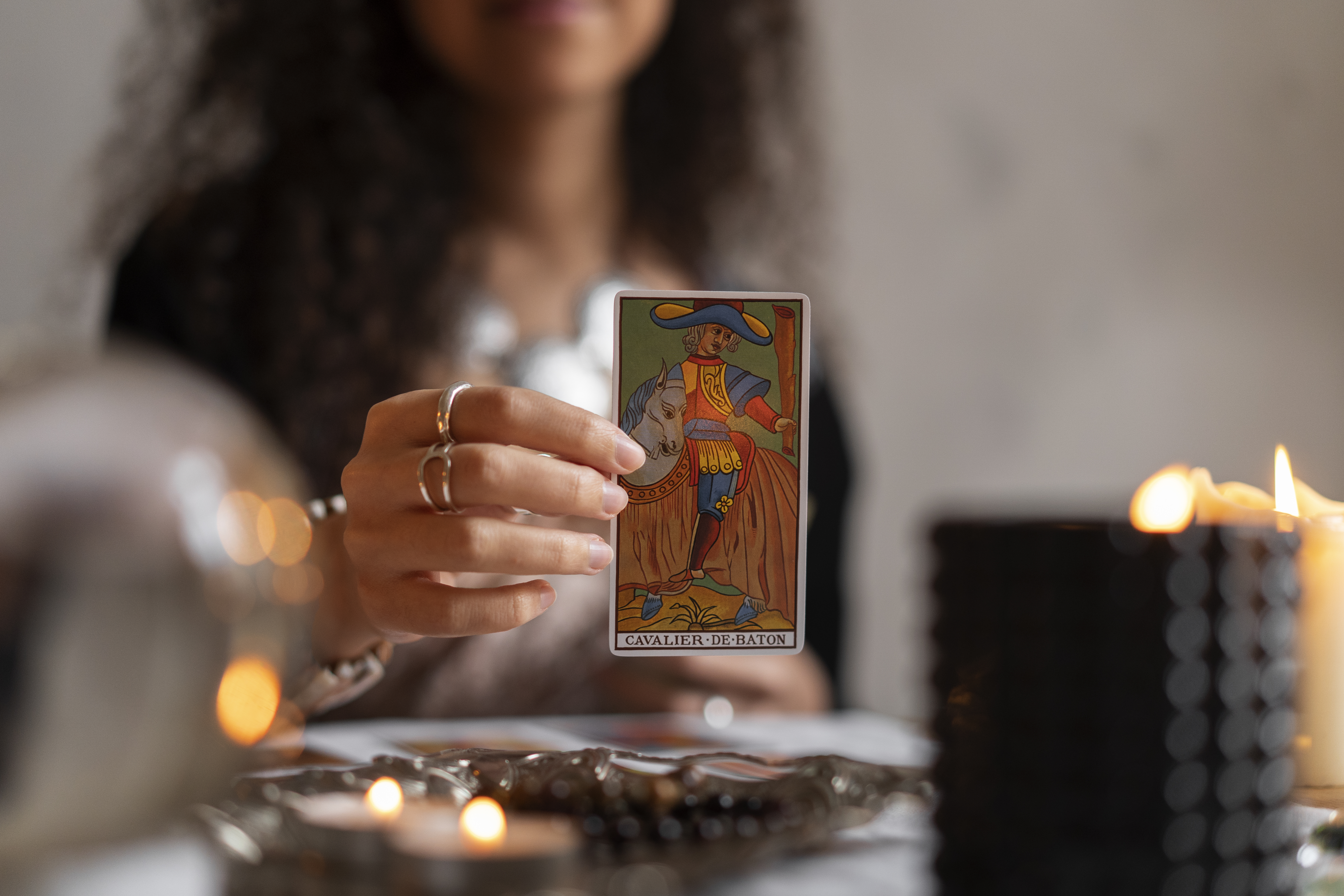 Mãos de uma mulher com anéis seguram uma carta de tarô Cavalier de Bâton com um cavaleiro em um cavalo, em um ambiente com velas acesas e objetos brilhantes