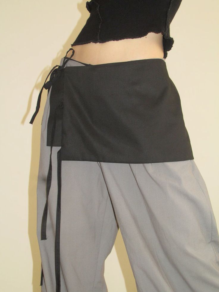 Pessoa de perfil, vestindo um top preto cropped e cal&ccedil;a cinza larga, com um avental preto amarrado na cintura, exibindo a barriga