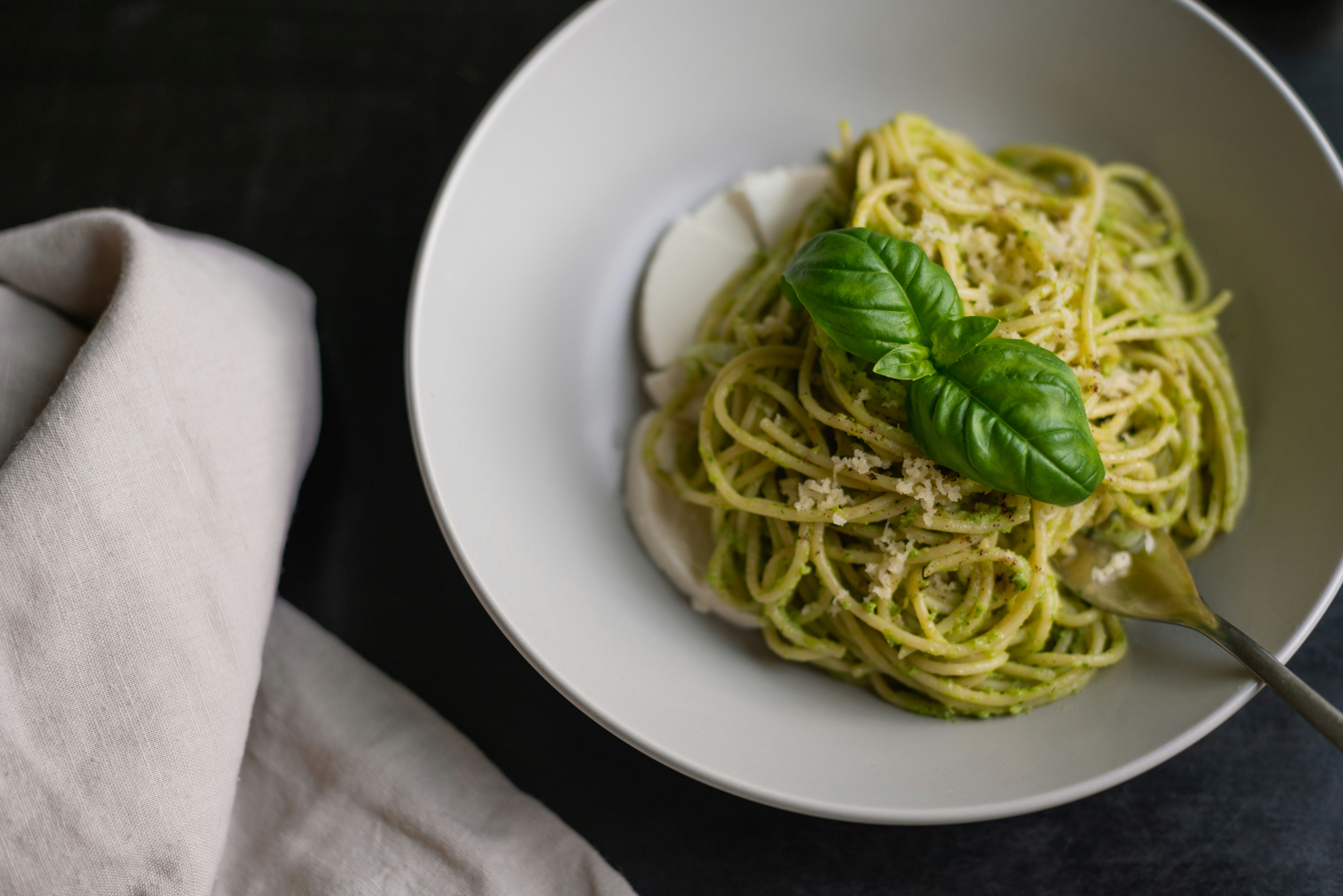 Prato de espaguete com molho pesto verde, queijo parmesão ralado e folhas de manjericão fresco, servido em uma tigela branca com fatias de queijo branco e um garfo, sobre uma superfície escura com um pano de linho claro