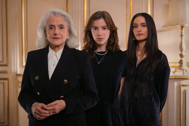 Três mulheres, uma idosa de cabelos brancos e duas jovens de cabelos castanhos, posam sérias em um ambiente luxuoso com paredes claras e detalhes dourados. A idosa veste blazer preto e camisa branca, as jovens usam roupas escuras e colares. Uma luminária clássica aparece ao fundo