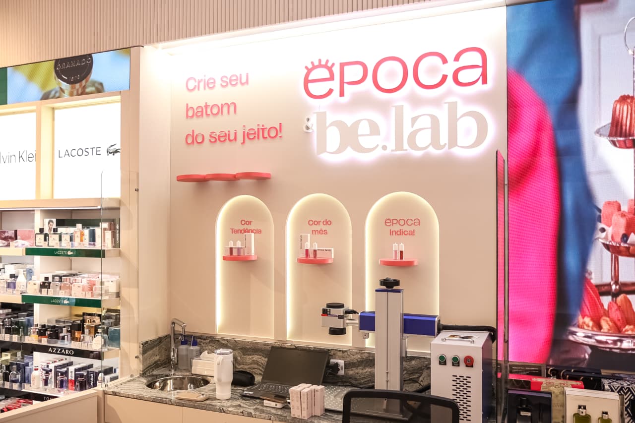 Ambiente de loja com balcão de mármore escuro e pia, exibindo um laptop, produtos de maquiagem e um aparelho de gravação a laser. Ao fundo, um painel luminoso com Crie seu batom do seu jeito! e Época be.lab, além de três nichos iluminados com batons e os dizeres Cor Tendência, Cor do mês e Época Indica!. À esquerda, prateleiras com perfumes de marcas como Calvin Klein, Lacoste e Azzaro. À direita, um painel digital exibe imagens coloridas