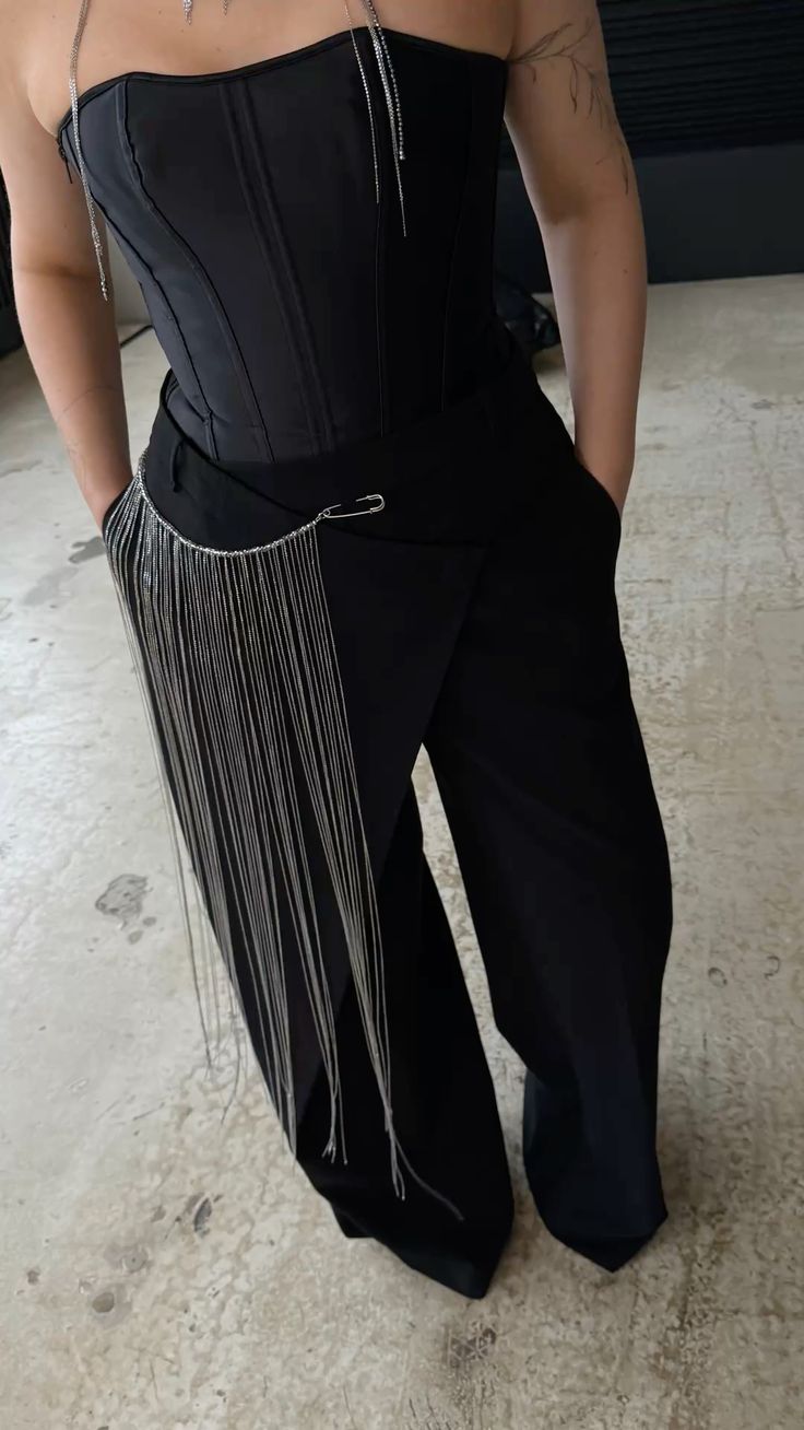 Pessoa de p&eacute;, vestindo um espartilho preto tomara que caia e cal&ccedil;as pretas largas. Um cinto de correntes prateadas com um alfinete de seguran&ccedil;a decora a cintura, com as m&atilde;os nos bolsos. O bra&ccedil;o direito tem uma tatuagem de galho. O fundo &eacute; um piso de cimento claro