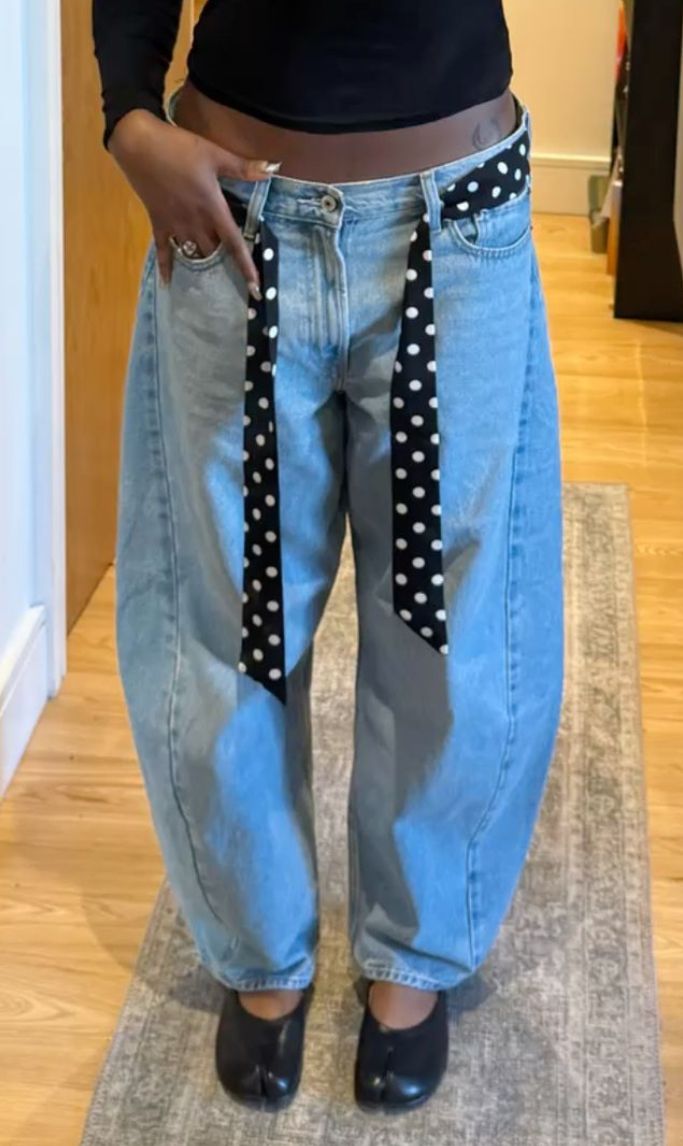Pessoa de pele escura vestindo cal&ccedil;a jeans larga de lavagem clara, top cropped preto e um cinto preto de bolinhas brancas. Nos p&eacute;s, sapatos pretos estilo tabi. A pessoa est&aacute; em p&eacute; sobre um tapete cinza claro, com piso de madeira ao redor