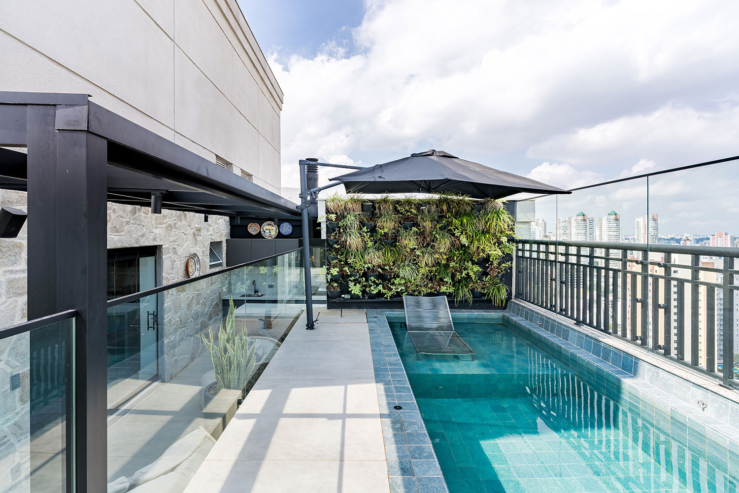 Cobertura com piscina retangular de azulejos azuis, espregui&ccedil;adeira submersa, parede verde e guarda-sol preto. &Agrave; esquerda, uma varanda com guarda-corpo de vidro e uma planta alta. Ao fundo, pr&eacute;dios da cidade sob c&eacute;u azul com nuvens