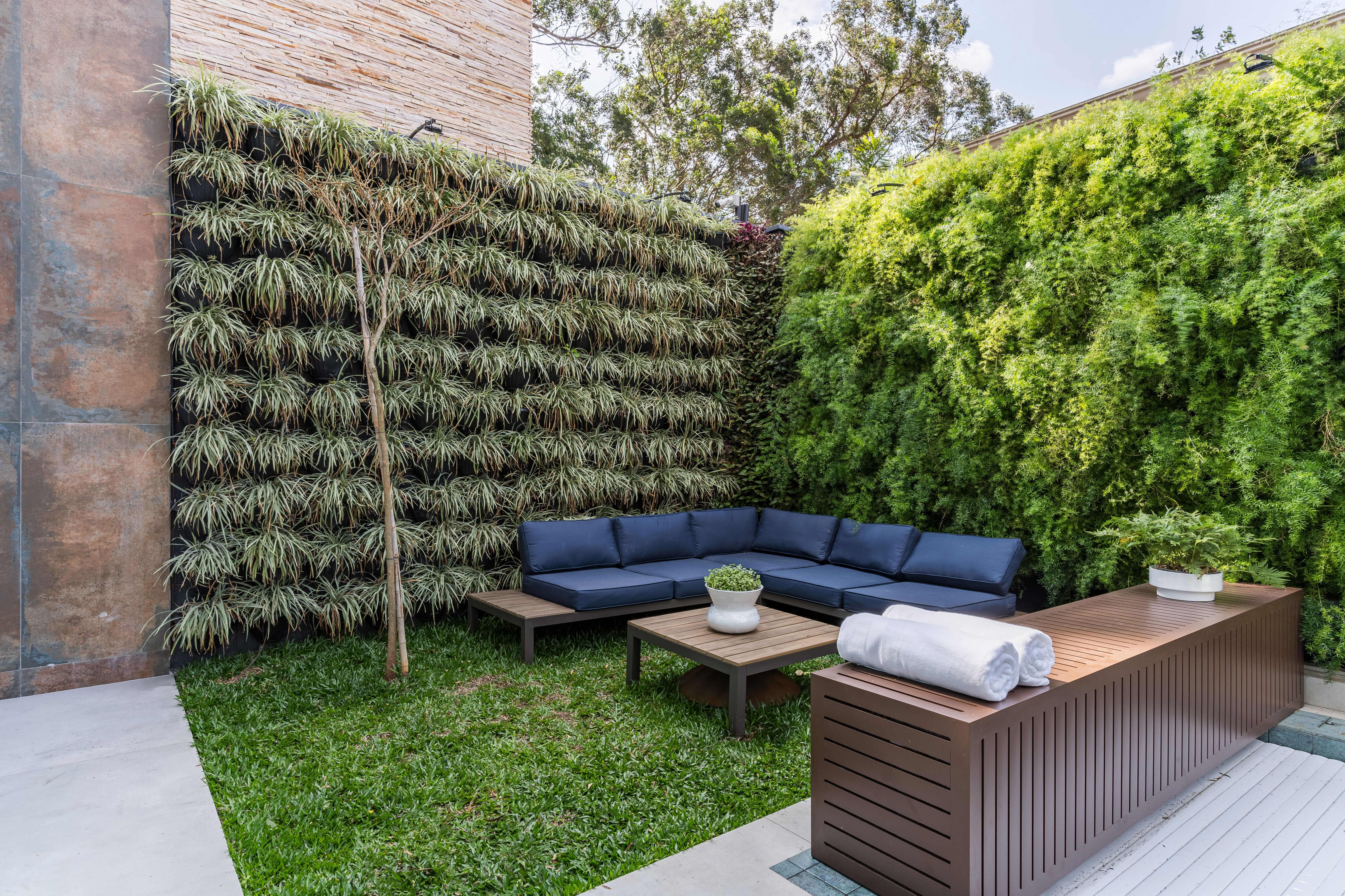 &Aacute;rea externa de uma resid&ecirc;ncia com jardim integrado e paisagismo abundante. O espa&ccedil;o &eacute; cercado por paredes revestidas de vegeta&ccedil;&atilde;o, com diferentes esp&eacute;cies de plantas criando um ambiente verde e intimista. Ao centro, um sof&aacute; modular azul-escuro forma uma &aacute;rea de estar, acompanhado por mesas baixas de madeira. Em primeiro plano, um grande banco ou aparador de madeira acomoda toalhas brancas enroladas e vasos de plantas. O piso combina &aacute;reas gramadas com revestimentos claros, enquanto as &aacute;rvores e o c&eacute;u ao fundo completam a composi&ccedil;&atilde;o
