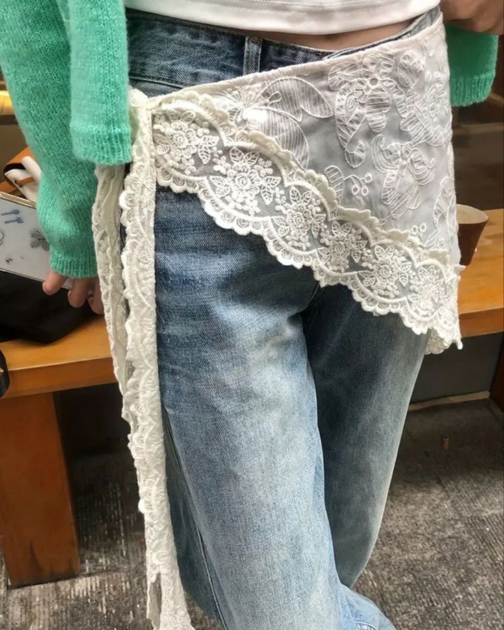Pessoa vestindo cal&ccedil;a jeans azul clara e um su&eacute;ter verde menta, com um cinto de renda branca floral amarrado na cintura, caindo sobre a perna esquerda