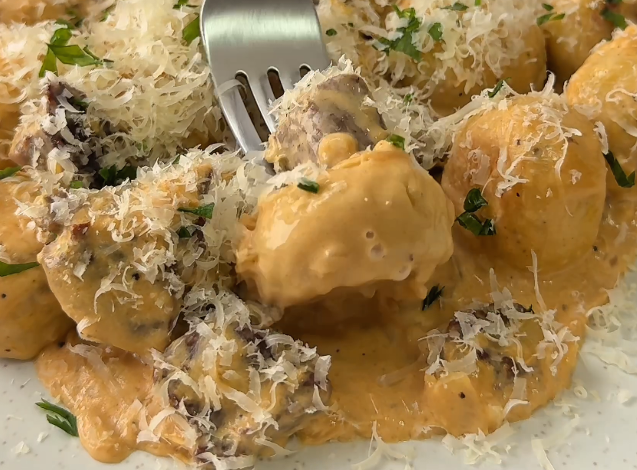 Garfo de metal prateado espetando um nhoque coberto por molho cremoso laranja, queijo parmesão ralado e ervas picadas, em um prato branco