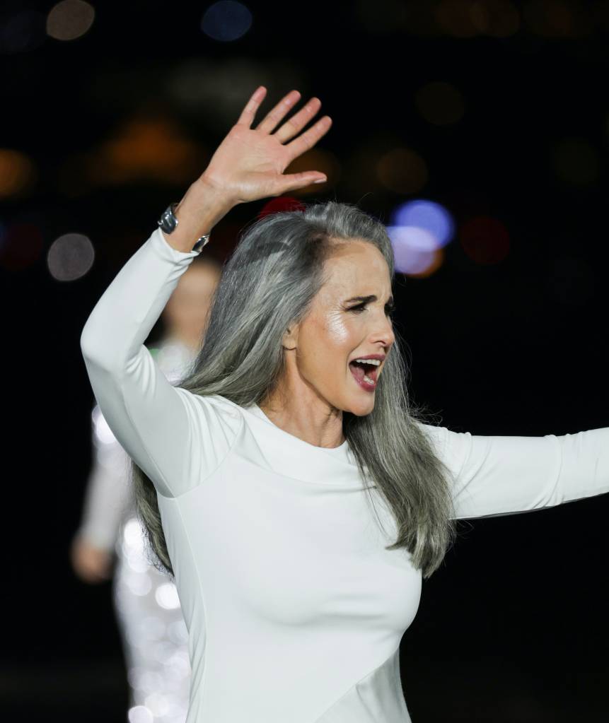 Mulher de cabelos grisalhos, vestindo um longo vestido branco, sorri com a boca aberta e levanta a m&atilde;o direita em um gesto de aceno, em um palco escuro com luzes desfocadas ao fundo