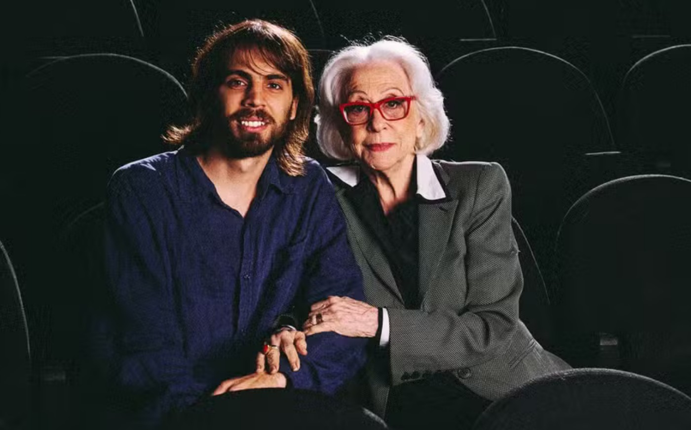 Um homem jovem de barba e cabelos longos sorri ao lado de uma mulher idosa de cabelos brancos e óculos vermelhos, ambos sentados em poltronas de cinema escuras