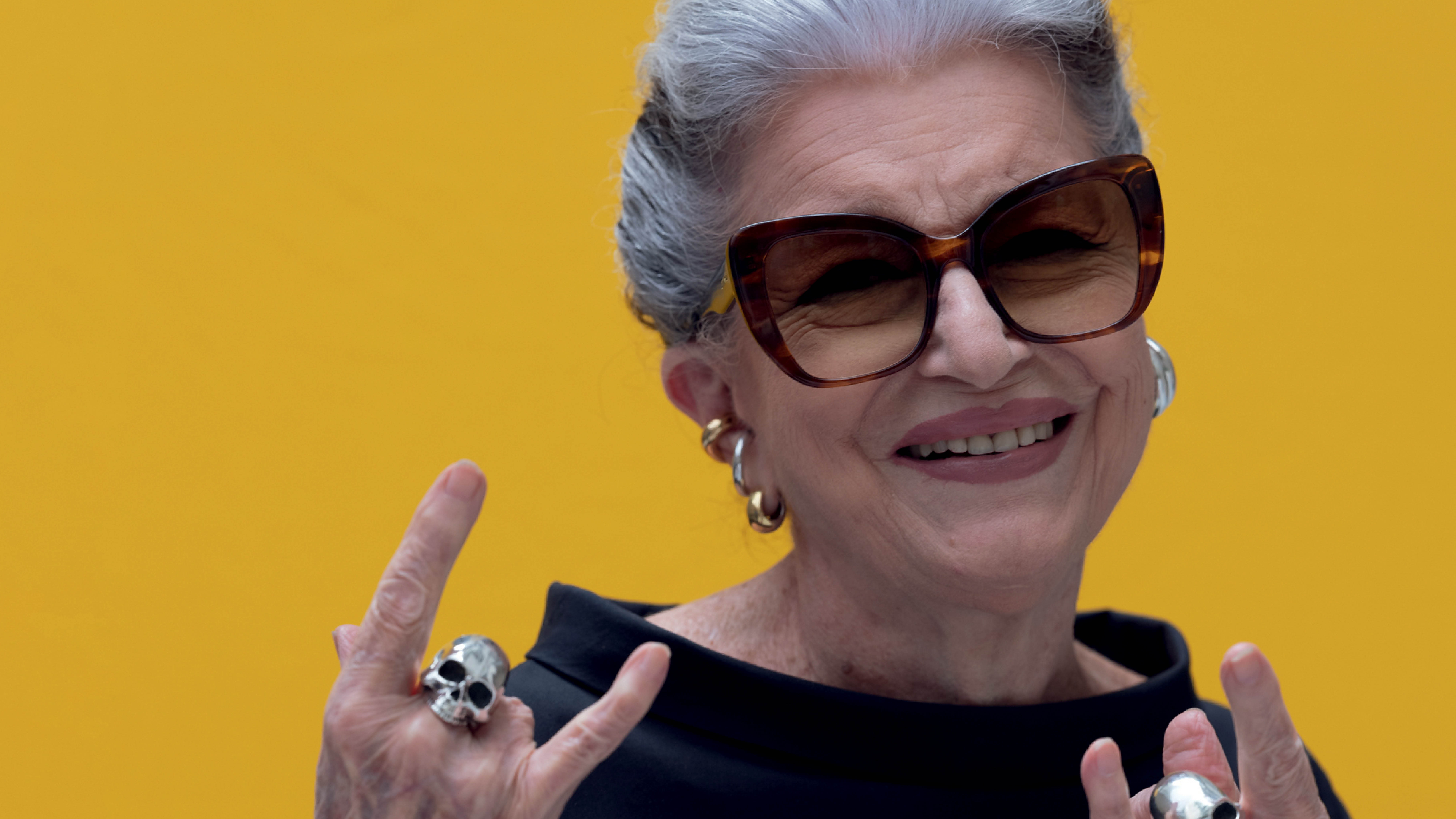 Mulher idosa de cabelos grisalhos, óculos de sol marrons e batom rosa, sorrindo e fazendo o sinal de chifres com as mãos, usando anéis grandes e brincos de argola, contra um fundo amarelo vibrante