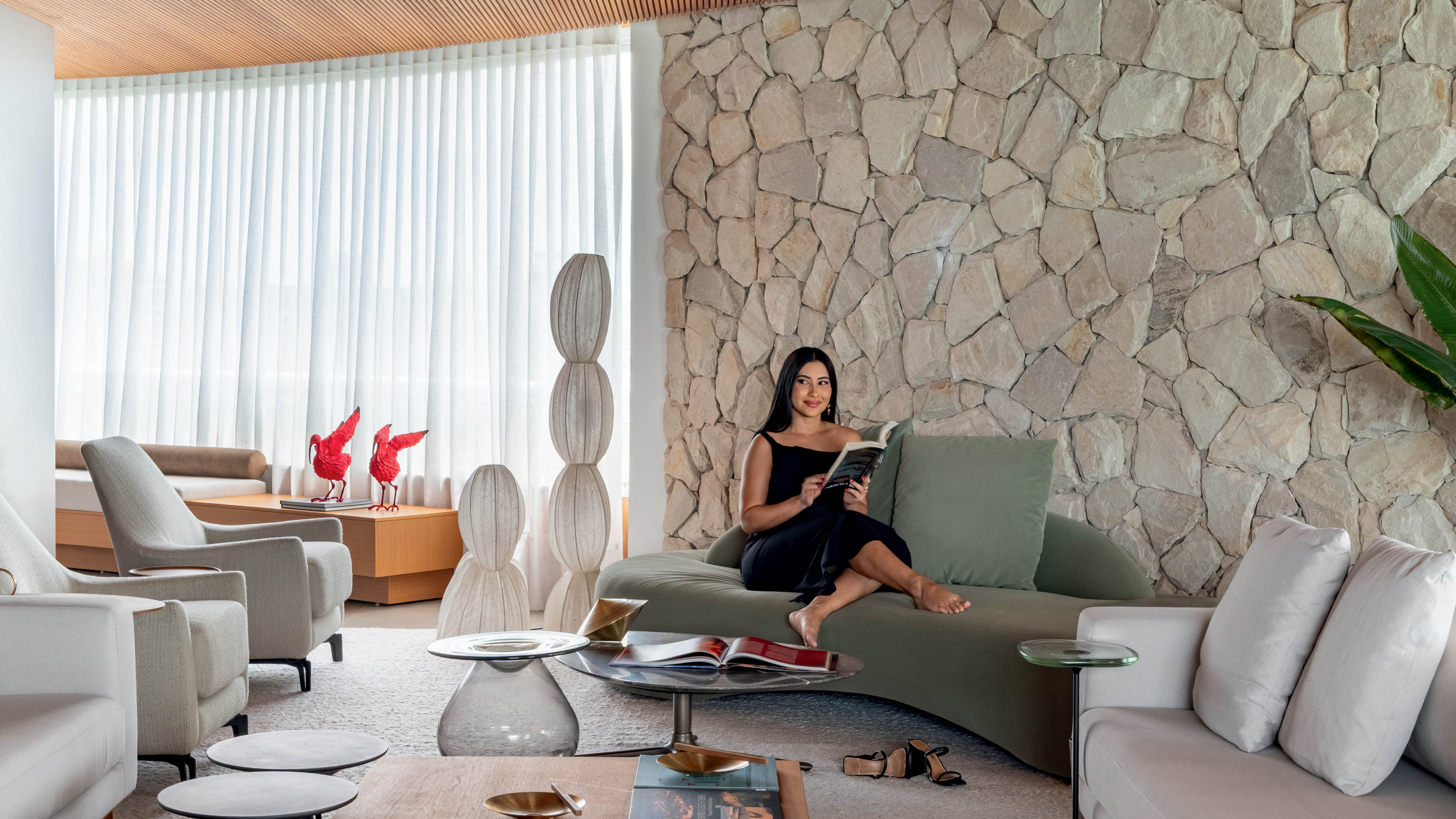 Mulher de cabelos longos e pretos, vestindo um elegante vestido preto, sentada descalça em um sofá verde-oliva, lendo um livro. Ela está em uma sala de estar moderna com uma parede de pedras claras, cortinas brancas translúcidas e um teto de madeira. Há poltronas claras, mesas de centro com livros e objetos decorativos, e esculturas brancas alongadas no ambiente