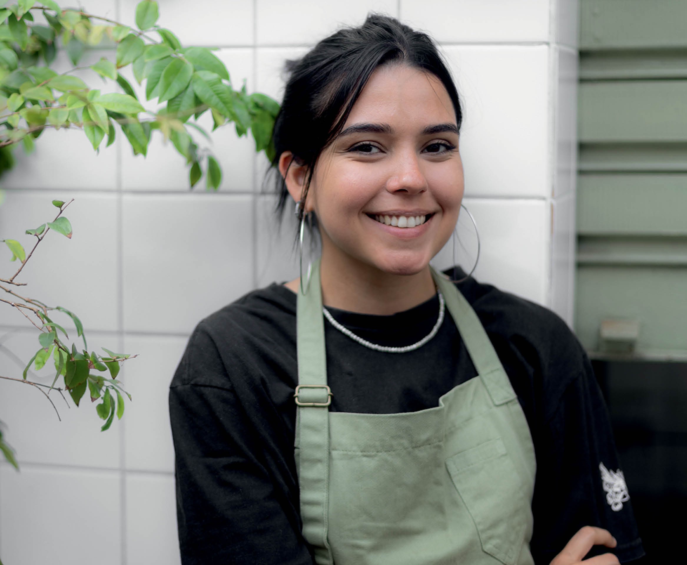 Jovem mulher de pele morena clara e cabelo escuro preso, sorrindo, usando brincos de argola grandes, colar de miçangas brancas, camiseta preta e avental verde-claro, em frente a uma parede de azulejos brancos com folhagens verdes à esquerda