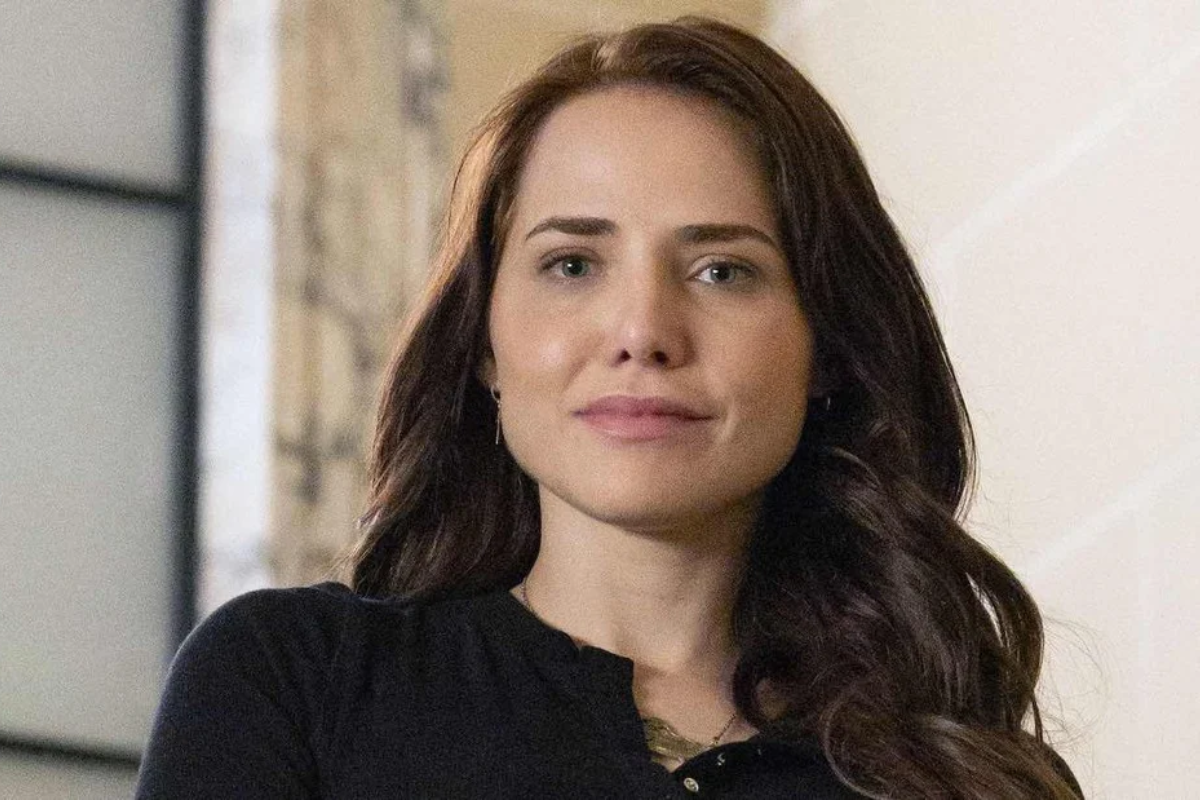 Mulher jovem de pele clara, cabelos castanhos longos e ondulados, olhos claros, vestindo blusa preta, sorri levemente para a câmera em ambiente interno com fundo claro
