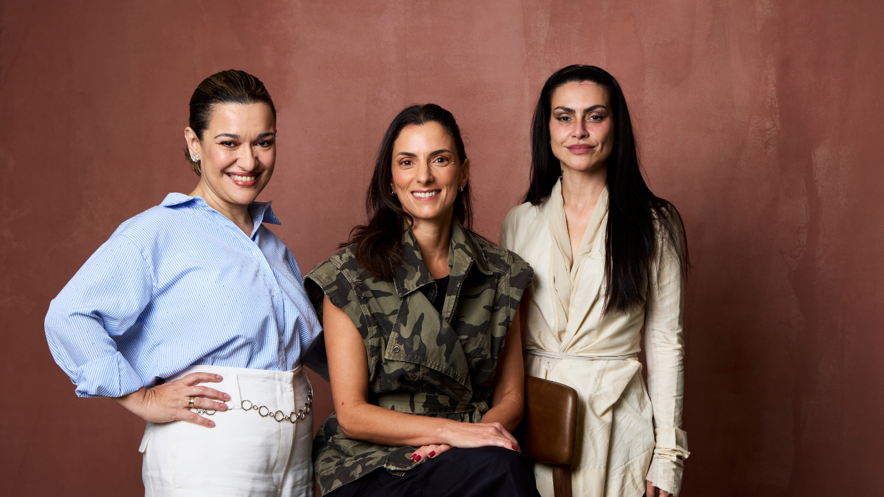 Três mulheres sorrindo, em pé e sentada, contra uma parede marrom. A da esquerda veste camisa azul listrada e calça branca com cinto de elos prateados. A do centro, sentada, usa colete camuflado e calça preta. A da direita, em pé, veste um casaco bege claro e tem cabelos longos e escuros