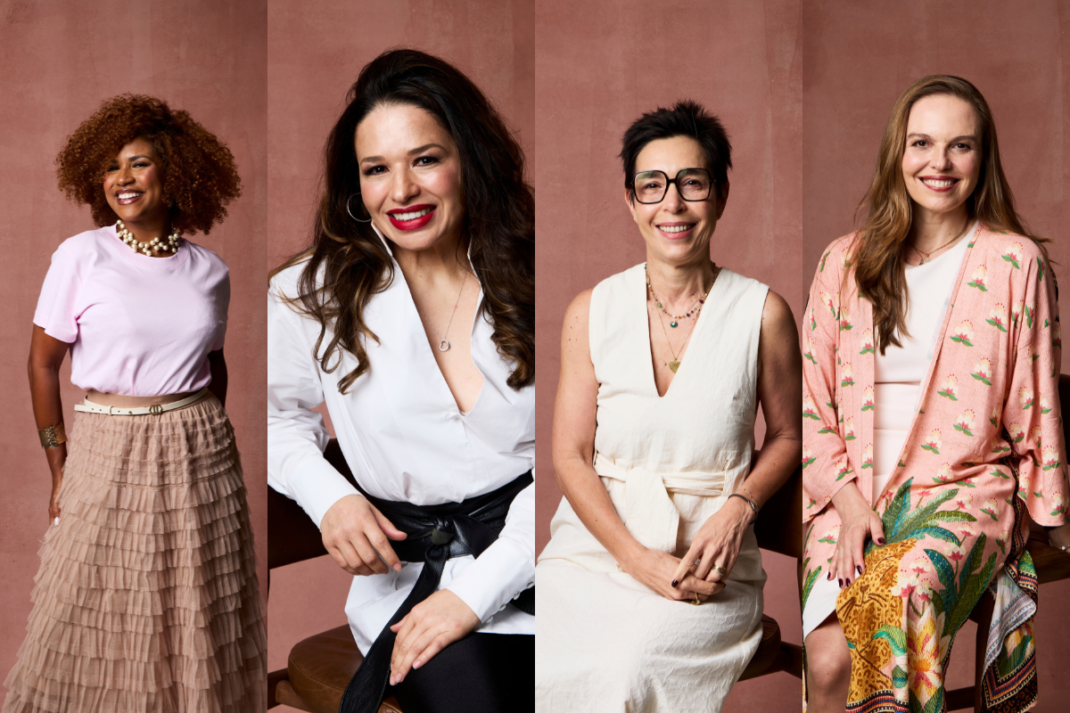 Quatro mulheres sorridentes, em fundo rosa. Da esquerda para a direita: mulher de cabelo cacheado ruivo, blusa rosa e saia bege; mulher de cabelo castanho escuro, blusa branca e cinto preto; mulher de cabelo curto escuro e óculos, vestido creme; mulher de cabelo castanho claro, quimono estampado rosa e verde