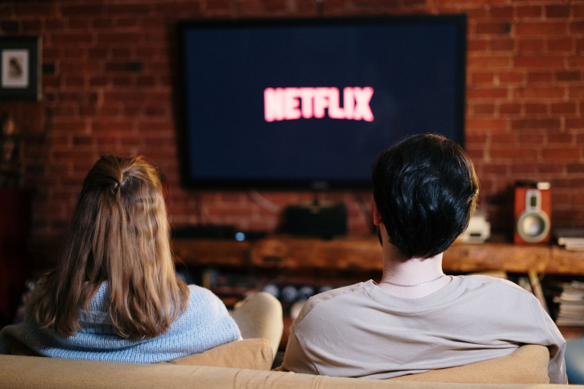 Um casal assiste à TV em um sofá marrom, de costas para a câmera. A mulher, à esquerda, tem cabelos loiros e usa um suéter azul claro. O homem, à direita, tem cabelos escuros e veste uma camiseta bege. Na televisão, o logotipo vermelho da Netflix aparece em tela preta. Ao fundo, uma parede de tijolos e uma estante com objetos.