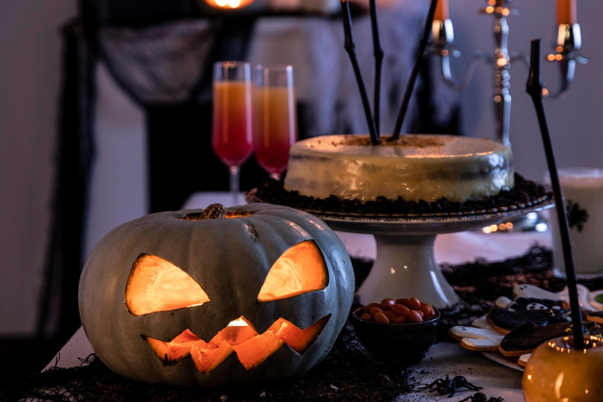 Mesa de Halloween com abóbora esculpida e iluminada, bolo em pedestal, dois copos de bebida laranja e vermelha, doces e aranhas decorativas, em ambiente escuro