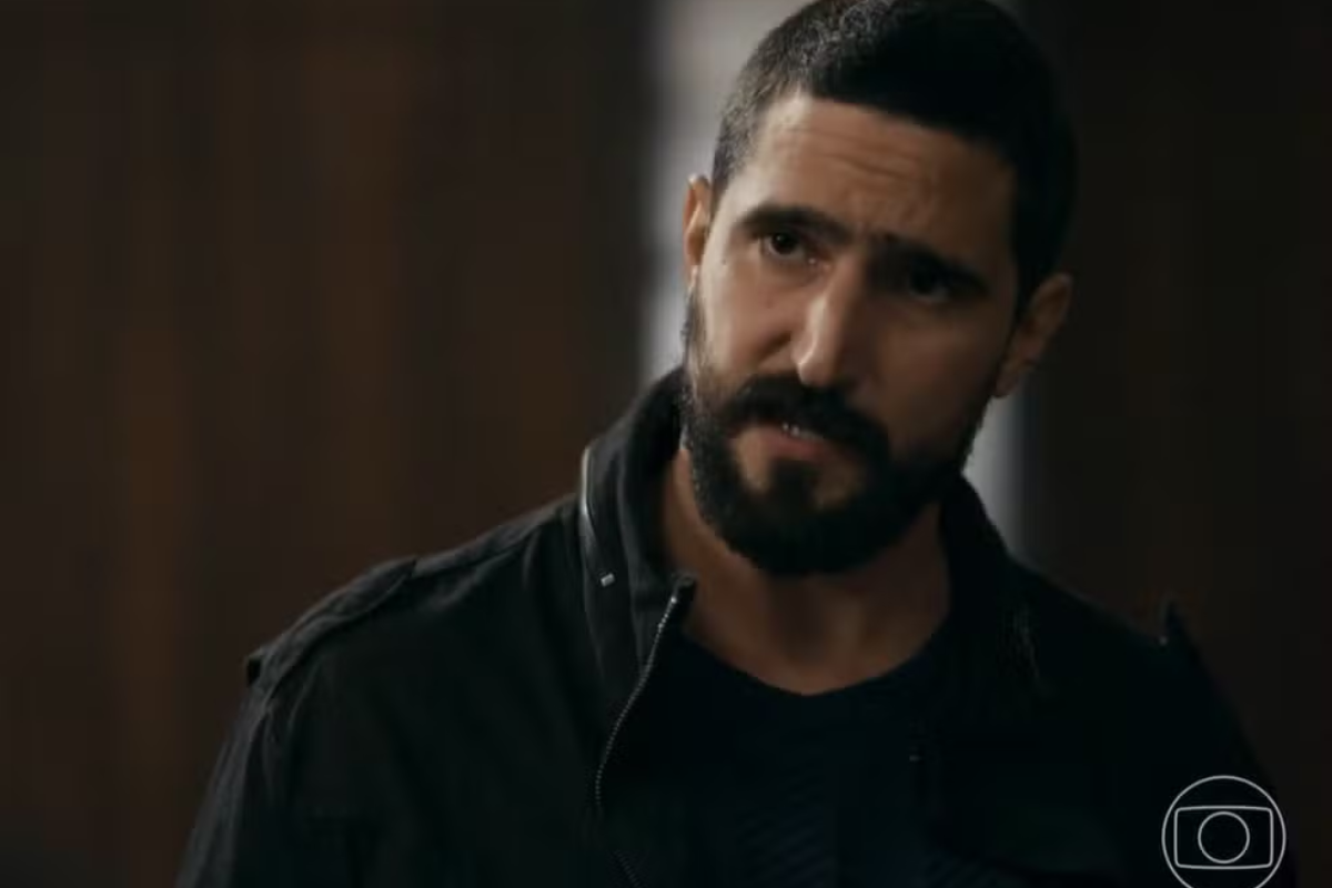 Homem branco de barba e cabelo escuros, vestindo jaqueta preta, olha para o lado com expressão séria e pensativa