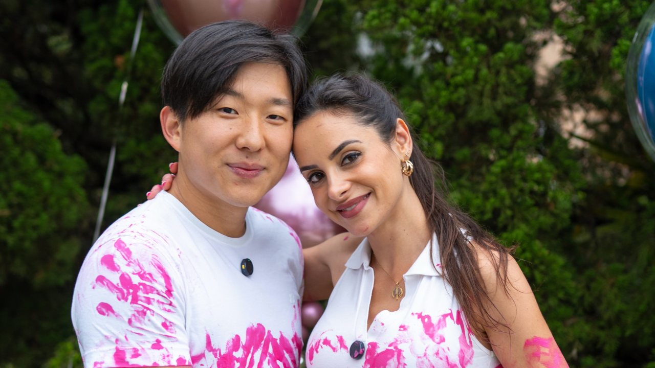 Um homem asiático e uma mulher de pele morena sorriem, com tinta rosa nas roupas brancas. Ele usa camiseta, ela uma blusa de gola com colar dourado e brincos de argola. Ambos têm tinta rosa nas roupas, e a mulher tem uma mão rosa no ombro do homem. Ao fundo, folhagem verde e balões coloridos.