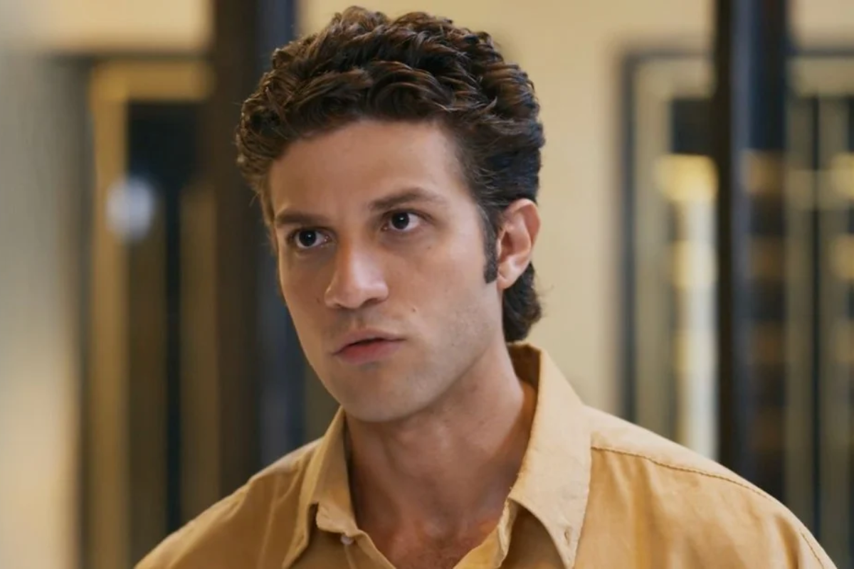 Homem jovem de pele clara, cabelo castanho escuro e ondulado, vestindo camisa bege, com expressão séria e olhar fixo para a frente