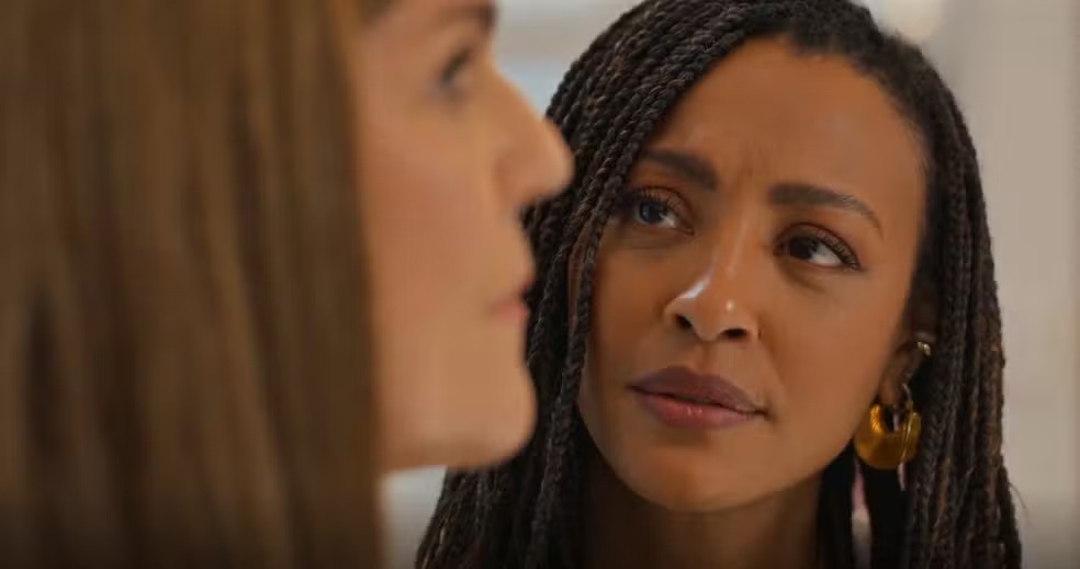 Uma mulher negra com tranças e brincos dourados olha atentamente para uma mulher branca de cabelos castanhos, que está de perfil e desfocada em primeiro plano