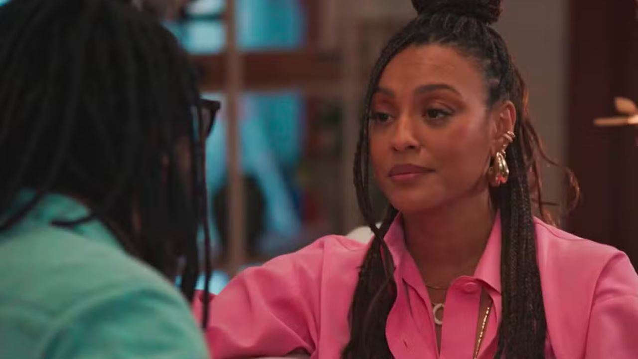 Mulher negra com tranças presas em coque, vestindo camisa rosa, brincos dourados e colar, olha atenta para um homem de dreads e óculos, de costas, em um ambiente interno
