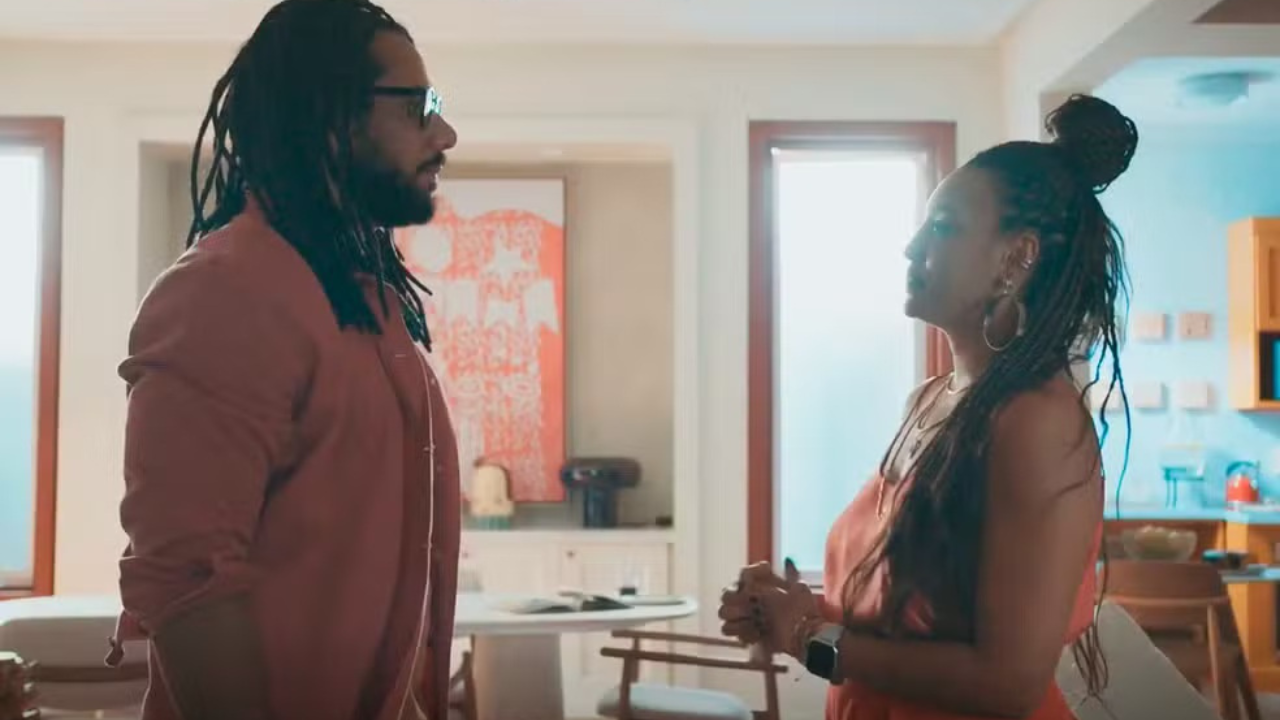 Um homem de dreads e óculos escuros, vestindo camisa rosa, conversa com uma mulher de tranças e blusa rosa, em uma sala com cozinha ao fundo