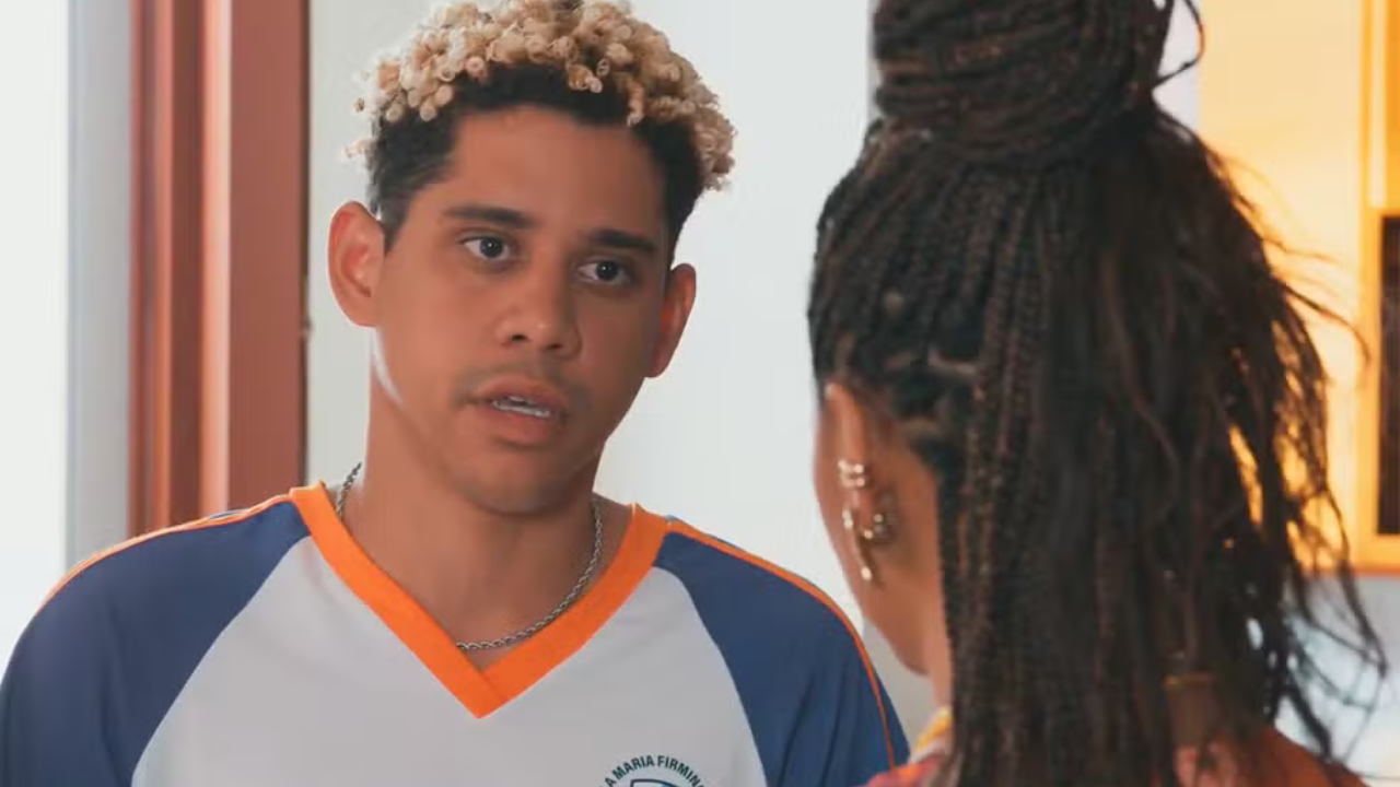 Um homem jovem, de pele clara e cabelos crespos loiros, usa uma camiseta branca com mangas azuis e gola laranja, olhando para uma mulher de costas com tranças escuras e brincos dourados