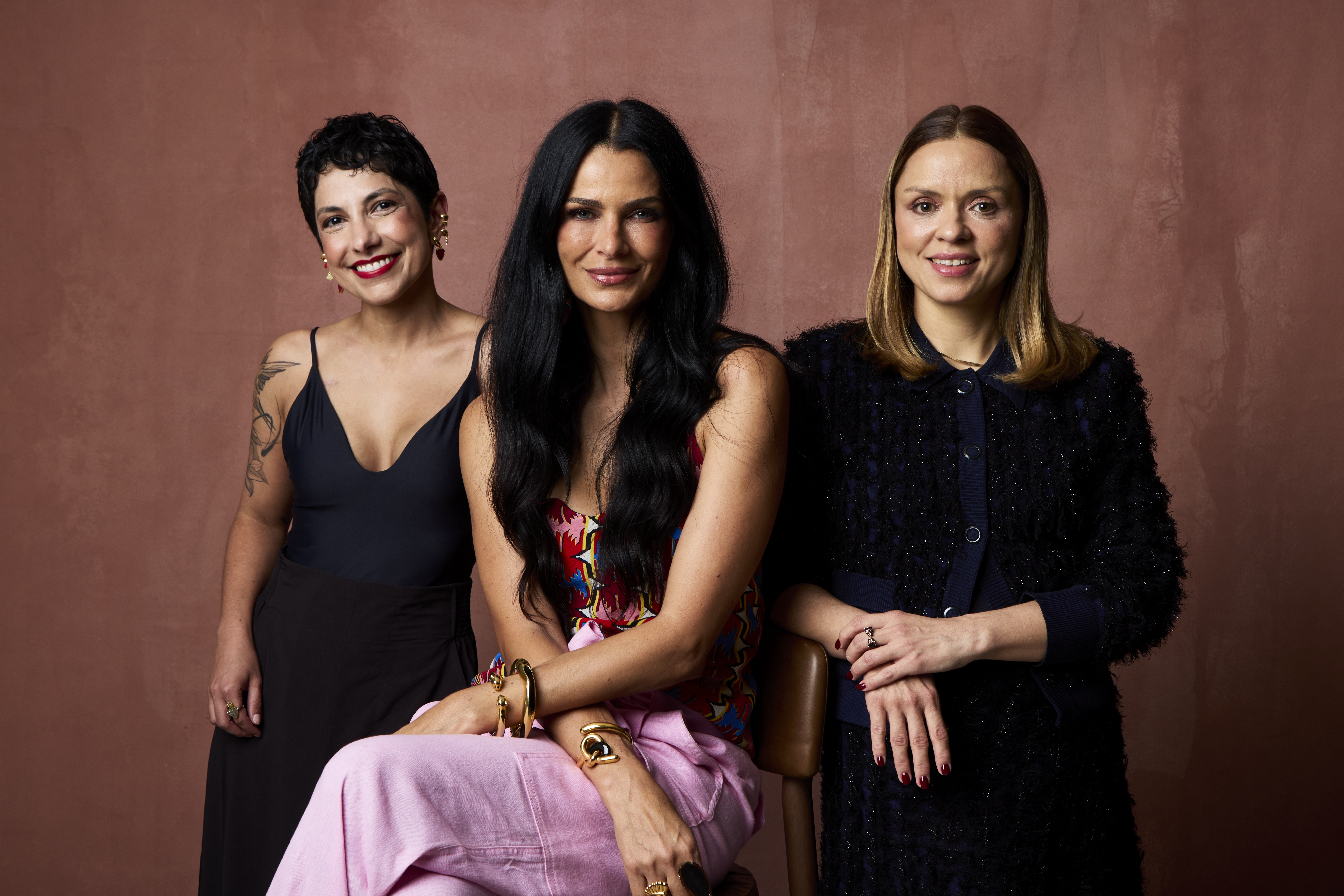 Três mulheres sorrindo para a câmera, com fundo marrom. A da esquerda tem cabelo curto, blusa preta e saia escura. A do centro tem cabelo longo preto, blusa colorida e calça rosa. A da direita tem cabelo loiro, casaco texturizado azul-marinho e saia escura