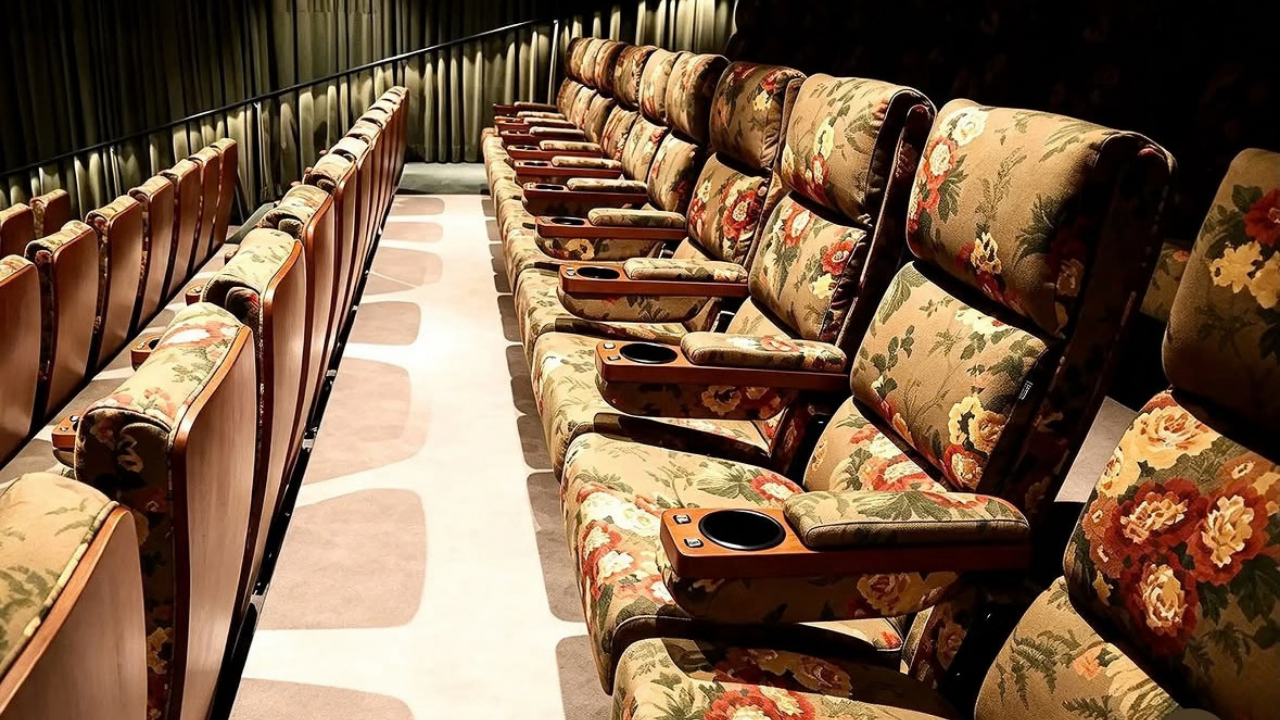 Fileiras de poltronas de cinema vazias, estofadas em tecido bege com estampa floral em tons de vermelho, amarelo e verde. Cada assento possui um apoio de braço de madeira com porta-copos preto. O corredor lateral tem um corrimão escuro e cortinas verticais ao fundo