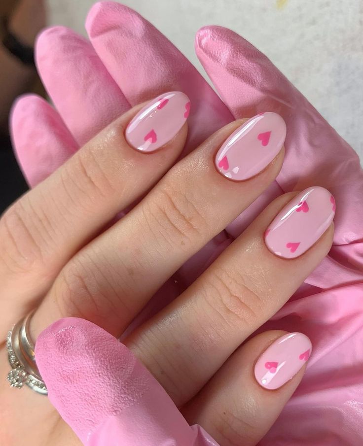 Mão feminina com unhas ovais pintadas de rosa claro e pequenos corações rosa choque, usando luva rosa na outra mão e anel prateado no dedo anelar
