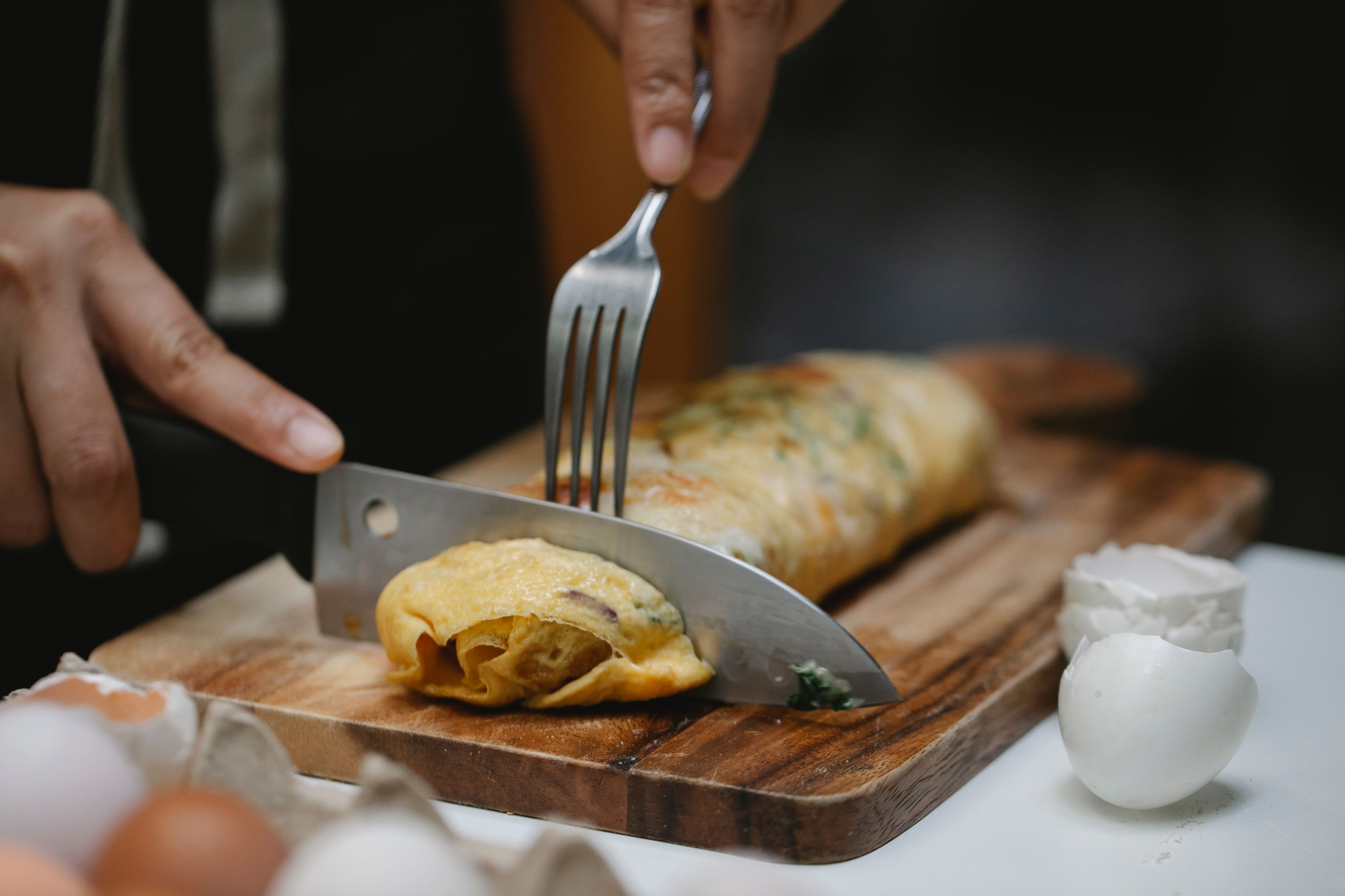 Mãos cortando um omelete enrolado com recheio verde e laranja em uma tábua de madeira, usando uma faca e um garfo. Cascas de ovos e ovos inteiros estão em primeiro plano