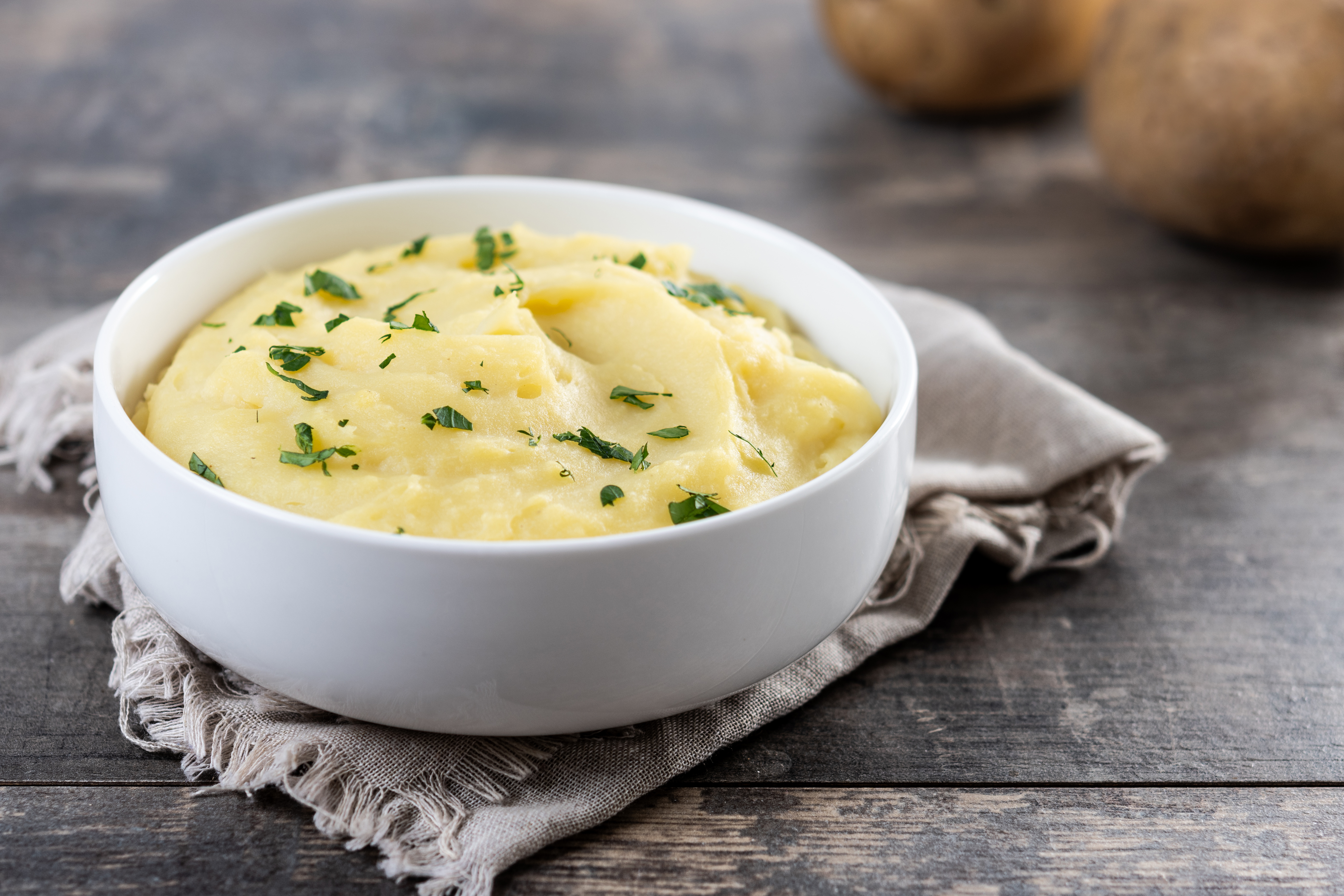 Purê de batatas cremoso em tigela branca, salpicado com salsa picada, sobre um pano de prato cinza e mesa de madeira rústica. Batatas inteiras desfocadas ao fundo