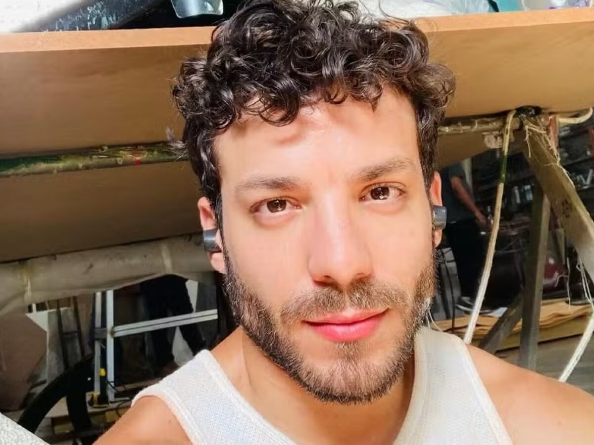Homem branco, de barba e cabelo cacheado escuro, sorri levemente para a câmera, usando regata branca e fones de ouvido pretos. Ao fundo, uma mesa de madeira e um ambiente de trabalho desorganizado