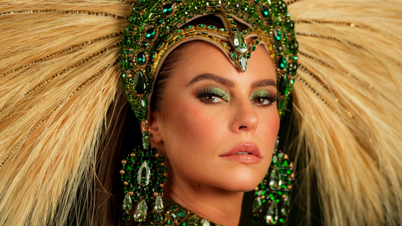 Mulher com maquiagem verde brilhante nos olhos e lábios rosados, usando um cocar e brincos grandes de pedras verdes e douradas, com plumas claras ao fundo