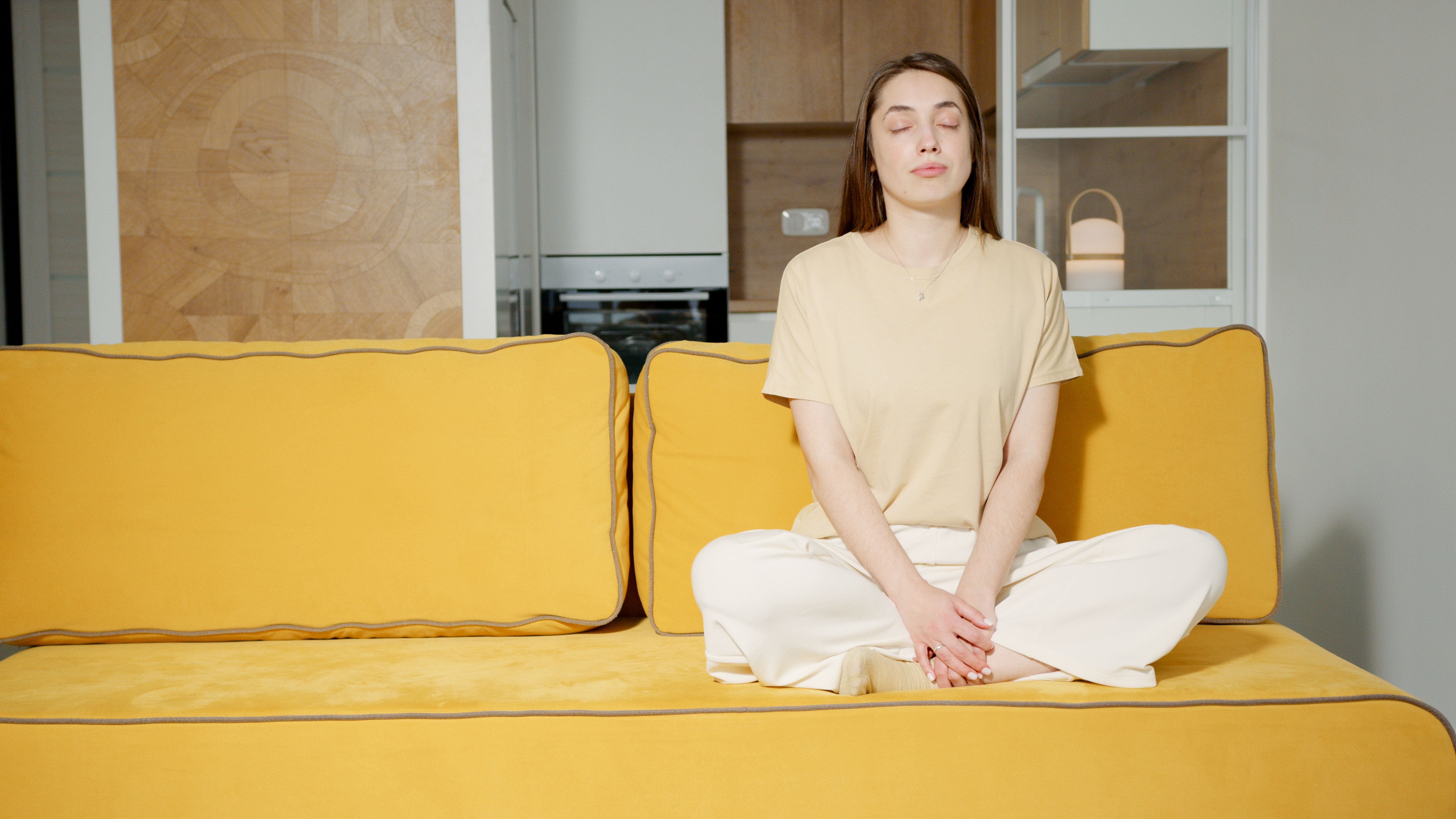 Mulher jovem de cabelos castanhos, camiseta bege e calças claras, sentada de pernas cruzadas em um sofá amarelo, meditando com os olhos fechados e mãos repousadas no colo, em uma sala com cozinha ao fundo