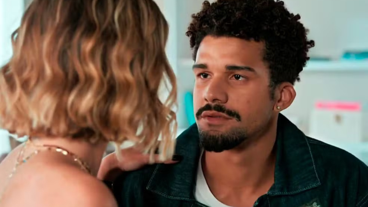 Um homem negro com cabelo crespo, barba e bigode, usando jaqueta jeans escura e camiseta branca, olha para uma mulher loira de costas, que usa um colar dourado. A mulher tem a mão no ombro do homem, que parece pensativo.
