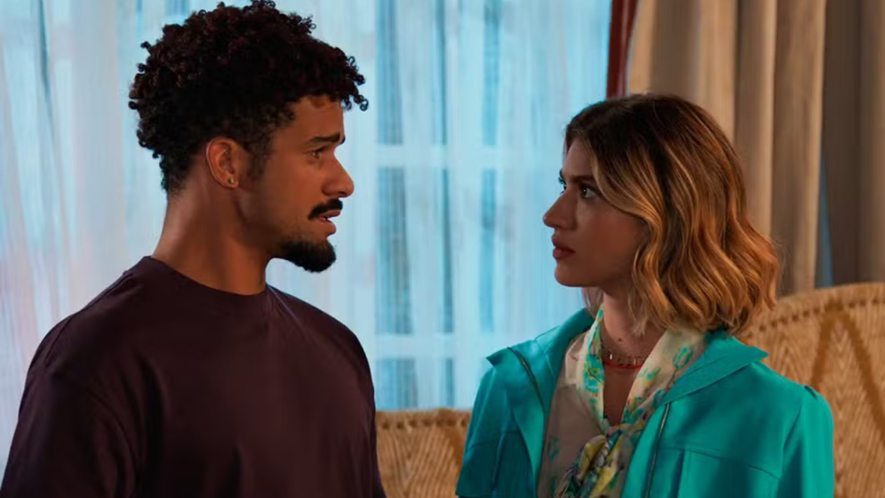 Um homem negro de cabelo crespo e barba, vestindo camiseta vinho, olha surpreso para uma mulher branca de cabelo loiro ondulado, que usa blazer azul-turquesa sobre blusa estampada e colar vermelho, com expressão séria. Eles estão em um ambiente interno, com cortinas claras ao fundo