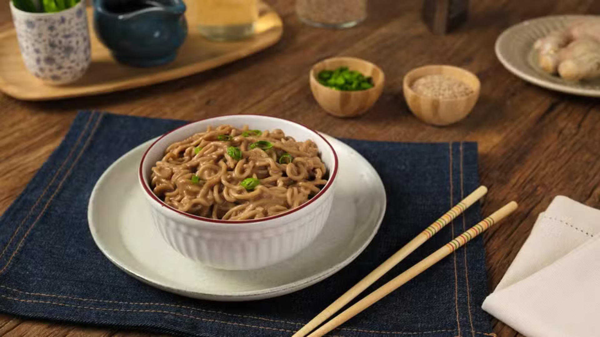 Tigela branca com macarrão oriental coberto por molho cremoso e cebolinha picada, sobre um prato branco e um jogo americano azul-marinho. Ao lado, hashis de madeira e um guardanapo branco. Ao fundo, potes com temperos e gengibre em uma mesa de madeira.