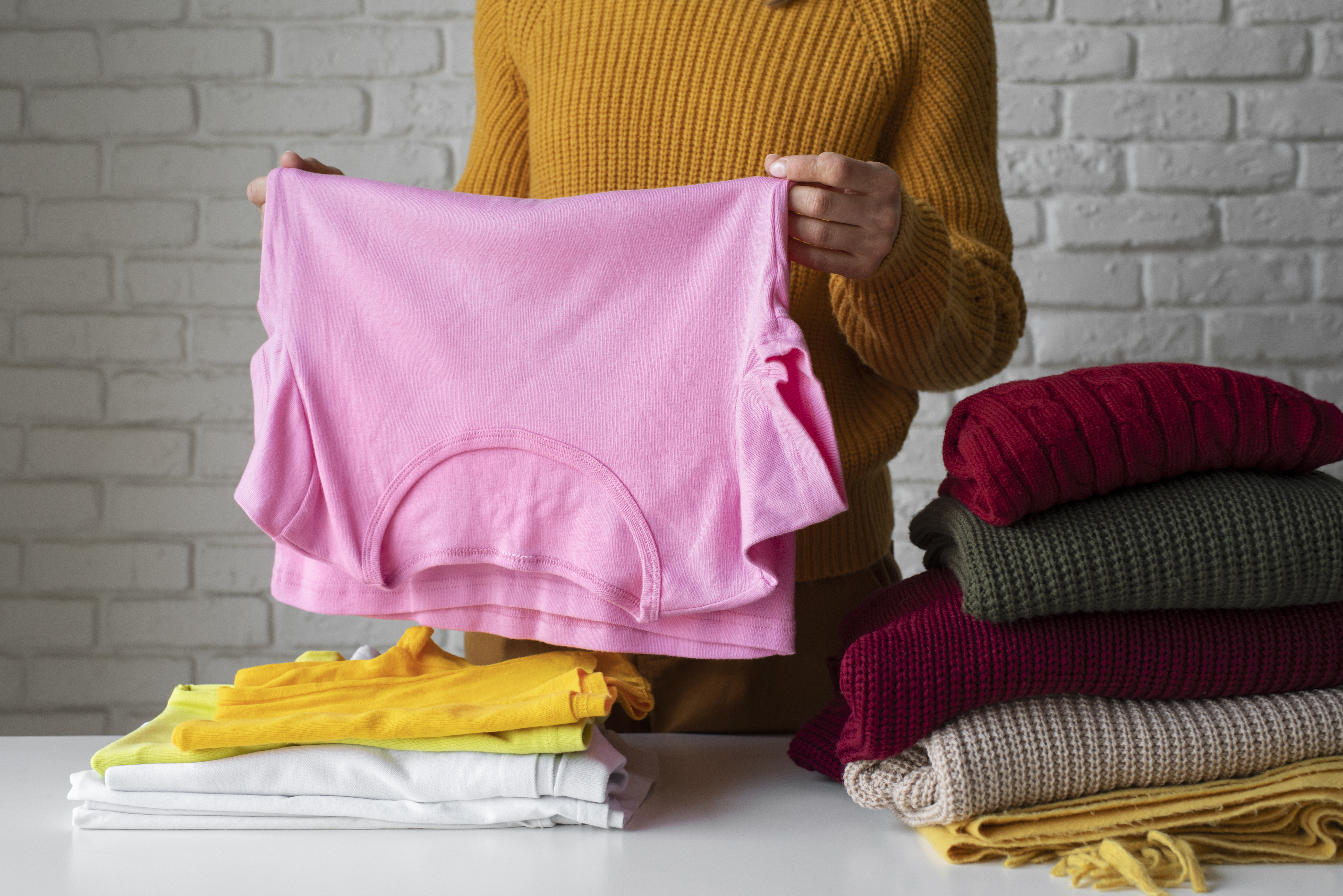Pessoa de suéter mostarda segurando uma camiseta rosa dobrada, com pilhas de roupas coloridas ao lado sobre uma mesa branca e parede de tijolos cinzas ao fundo